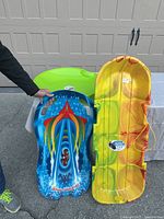 Three colorful plastic snow glider sleds standing vertically against a garage door: green, blue with flame design labeled Snow Skimmer, and yellow with mounted pull rope.