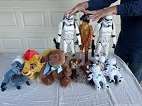 Lot photo showing various stuffed animals including Stitch, Simba, teddy bear, hedgehog, and two white bears, along with two Stormtrooper figures and one Star Wars pilot figure on a table.