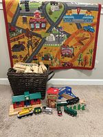 Full view showing woven basket filled with wooden track pieces, 7 colorful train cars arranged in front, small accessories, and play mat hanging on wall