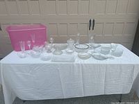 Assorted glass and crystal items displayed on a white cloth with garage door background. Includes various bowls, vases, footed dishes, and a pink plastic storage bin.