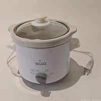 White Rival slow cooker from front angle with glass lid in place, attached power cord visible, dial control on front.