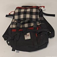 Front view of black and white plaid backpack with red zipper accents, showing adjustable straps and front flap with buckle closures.