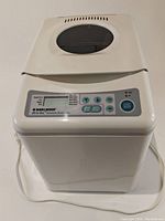 Front and side view of the breadmaker showing the control panel, viewing window in the lid, and power cord.