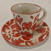 Front view of hand-painted clay espresso cup on matching saucer, showing red floral pattern and rustic crackle glaze.
