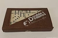 Closed brown faux leather case with clear plastic window showing white domino tiles with black pips inside. Case labeled 'Double 6 Dominos B.J.C.'