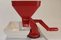 Front angled view of the red manual tomato press showing the funnel hopper, red crank handle, white catch container, and suction base attached to the bottom.