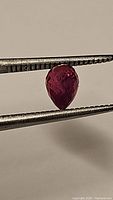 Close-up photo of a fancy briolette cut pink-purple sapphire held by tweezers, showing facets and inclusions.