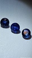 Three round denim blue natural sapphires displayed side by side on a white textured background, highlighting their round faceted cuts and blue color.