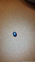 Oval cabochon natural blue star sapphire gemstone placed on textured brown surface, displaying distinct 6-ray star asterism effect under light.