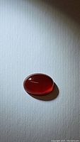 Oval cabochon natural fire red carnelian gemstone placed on a white surface with light and shadow showing translucence and rich red color.