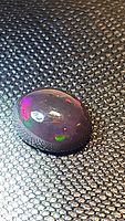 Natural oval cabochon black opal on textured surface showing bright multicolor flashes, primarily red and green colors.
