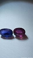 Photo of two natural oval cut sapphires, blue and pink, shown side by side on a white surface under natural lighting.