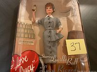 Front view of Barbie doll in original display box featuring 'I Love Lucy' branding and 'Vita Meat Vegamin' bottles backdrop. Doll holds a small bottle and wears gray checkered dress.