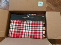 Top-down view of an open cardboard box displaying a folded red, white, and green plaid tablecloth labeled Robert Stanley Boat Collection.