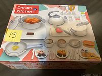 Photo of the Dream Kitchen toy set box showing contents including pot with drumstick, kettle, pans, utensils, and play food