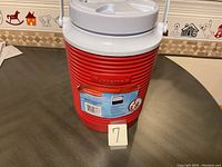 Red Rubbermaid water jug with white lid and carrying handle, showing whole jug and sticker label.