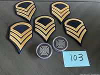 Top view of five embroidered military sergeant rank patches in gold thread on black fabric backing, arranged around two circular sterling badges with a cross motif inside clear plastic protective cases.