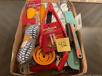 Top view of cardboard box containing new kitchen utensils and tools including spatulas, tongs with thermometer, citrus juicer, potato masher and kitchen towel.