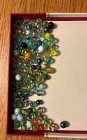 Top-down photo of a collection of 73 assorted glass marbles in a box with a red rim.