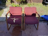 Front view of two chairs with wooden arms and legs, maroon fabric upholstery with visible tears on one seat
