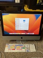 Front view of Apple's 21.5-inch iMac powered on showing the Ventura OS desktop, USB keyboard with colored key markings, and mouse in front. Shows general physical condition of computer and peripherals.