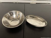 Photo showing round deep metal bowl and oval shallow metal bowl on a black surface.