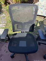 Front view of black mesh office chair placed outdoors on concrete and grass, showing mesh fabric on seat and backrest, adjustable armrests, and wheeled base.