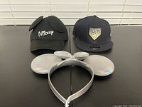 Photo of 3 headwear items on table, showing front views: black Mickey Mouse cap with ears, Milwaukee Brewers fitted hat, and silver holographic Minnie Mouse ears headband.