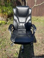 Front view of black leather office chair placed outdoors on grass with wooden fence and plants in background.