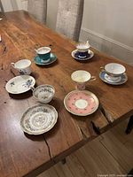 Seven antique teacups and saucers arranged on a wooden table, showing variety of floral and ornate designs in blue, pink, white, turquoise with gold trim.