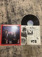 Vinyl record album with cover and newspaper-style insert on table, showing artwork and items.