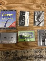 Six cassette tapes arranged on a wooden surface including Stephen R. Covey's audiobook, black and white tape cases, and a green Dinosaur Jr. tape.