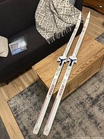 Full length view of the pair of white junior cross country skis laid on a wooden table with visible red branding and blue/white bindings.