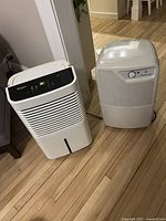 Two portable dehumidifiers side by side on a wood floor near power outlet.