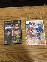 Two video games side by side on a wooden surface: PlayStation 2 'WWE SmackDown! Here Comes the Pain' and Wii 'Mario & Sonic at the Olympic Games'.