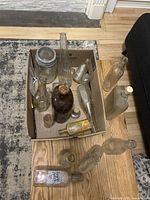 Several vintage glass bottles of varied size and shape inside cardboard box and on floor