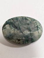 Oval polished emerald gemstone weighing approximately 55 carats, showing natural green shades and black veining.