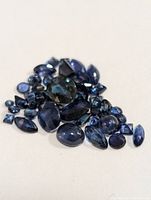 Multiple loose blue sapphire gemstones shown in various cuts including oval, pear, marquise, and round. Deep blue colored stones laid out on a white background.