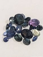 Photo showing a collection of assorted sapphire gemstones in various cuts and colors including blue, teal, yellow, and purple, arranged loosely on a plain background.