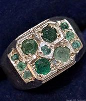 Close-up top view of silver ring showing ten round emerald stones embedded in rectangular layout.