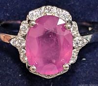 Close-up front view of silver ring with large faceted oval pink ruby center stone and surrounding clear CZ stones in halo style setting.