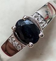 Close-up angled view of the silver ring showing the oval faceted dark blue sapphire center stone and small clear accent stones on either side.