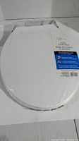 White plastic toilet seat inside slightly worn packaging, showing oval shape and intact cold press-wrap.