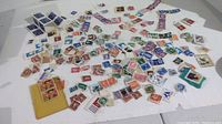 Wide image showing over 100 loose used postage stamps in various colors and designs scattered on a white surface.