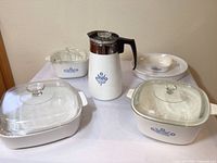 Overview of 3 CorningWare baking dishes with blue cornflower pattern, 2 all-white dishes, clear glass lids, and coffee carafe with blue cornflower pattern.