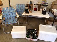 Two blue-striped folding chairs, two white Styrofoam coolers on floor, cardboard box holding multiple green propane bottles, table in background with lanterns and camping supplies.