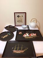 View of three Couroc black lacquer trays with inlaid designs including two sailing ships and one tiger, a framed Japanese woodblock print signed R. Boegehold 1930, a white ceramic teapot with woven handle, a small blue and white bowl, and a rectangular ruler on a white tablecloth.