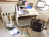 Wide angle showing assortment of canvases, frames, papers on a table and floor with photo album refills and foam spheres, including a standing paper cutter with magnifier lamp.