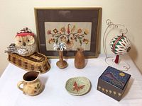 Overview of all items on white table including pottery, wood vases, framed art, woven bird, thread ball on stand, and decorative box.