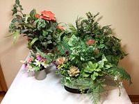 Wide view of all three faux greenery and floral arrangements together on a table against a beige wall.